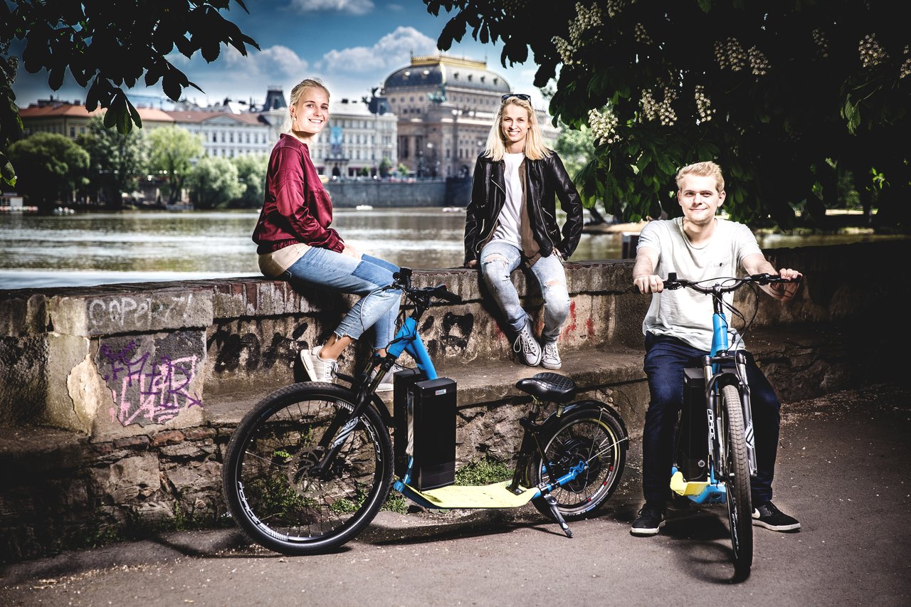 Two people sitting on a wall on the outskirts of Prague, with a man sitting on an electric scooter in front of them