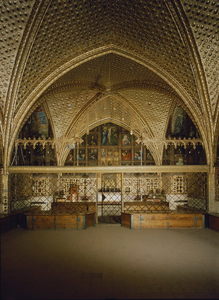 A Gothic arch entirely covered in gold and decorations, protected by a grille, with paintings in the background