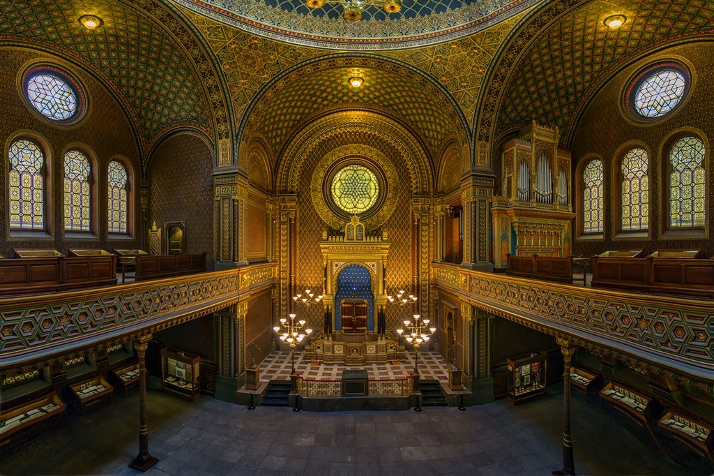 A majestic hall with walls carved in gold featuring Hispano-Moorish motifs, and circular stained-glass windows with designs