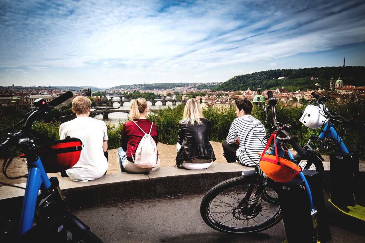 Four people sitting with their backs to the camera, gazing out at the city of Prague, with electric scooters behind them