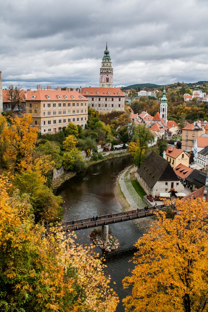 A dark river in the middle of a city with red-tiled roofs, dominated by a tall, ornate tower under a cloudy sky.